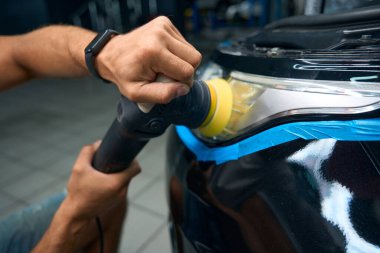 Guy with muscular arms cleans the headlights of a car, he works in a car repair shop