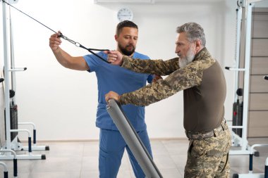 Military man works out on a special simulator during the recovery period, he is helped by an experienced trainer