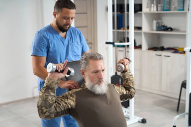 Military man in camouflage clothes works out in the gym in a rehabilitation center, an experienced physiotherapist works with him