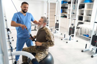 Rehabilitation specialist is working with a military patient in the gym, the patient is sitting on a fitball