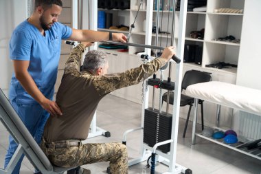Experienced rehabilitation therapist works with a patient in the gym, a specialist uses a special simulator