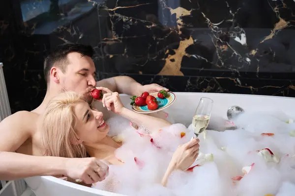 Female and a male feast on strawberries and champagne in a foam bath, a man gently hugs a woman
