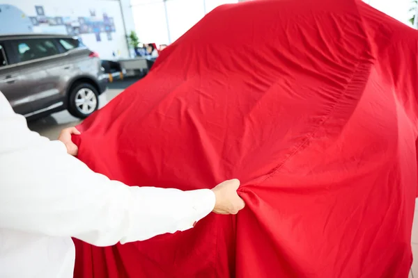 Man removes a red cover from a car, there is a large selection of cars in a car dealership