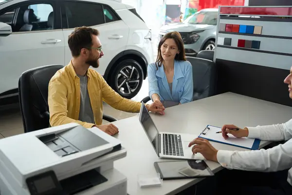 Spouses communicate with a manager in the office area of a car dealership, people sit at the table