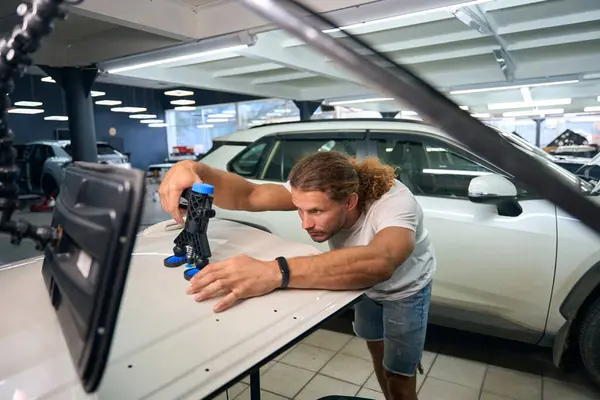 Long-haired guy uses a straightening pump at work, he is repairing a white car