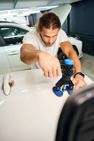 Long-haired guy is working on straightening a white car, he uses special gadgets