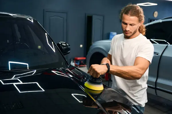 Guy in a car repair shop polishes the body of a black car, he uses a special device