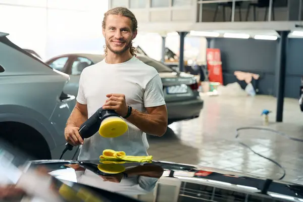 Smiling man works in a car repair shop, he polishes a car with a special grinder and a soft napkin