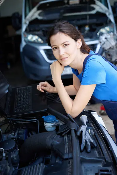 Woman auto mechanic uses a laptop for work, she is located under the open hood of a car