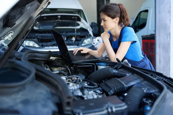 Female auto mechanic uses a laptop for work, she is located under the open hood of the car