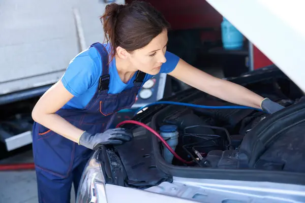 Cute female auto mechanic in work overalls works under the hood of a car, master in protective gloves