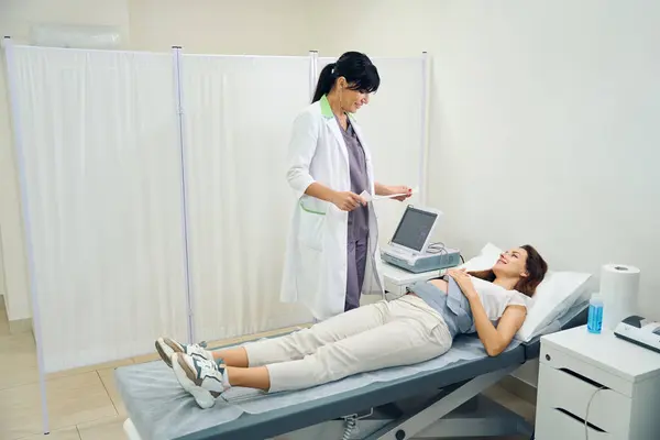 大人のヨーロッパの女性婦人科医は 診療所で若い白血病妊婦の腹の電気カルダイオグラムを見ています 妊娠検査の概念 — ストック写真