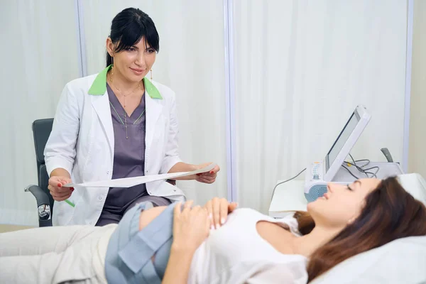 大人のヨーロッパ人女性婦人科医が若い白血病の妊婦の腹の電気カルダイオグラムを保持し 診療所で彼女を見ています 妊娠検査の概念 — ストック写真