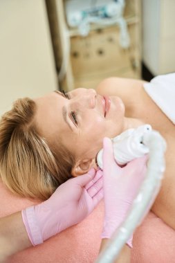 Bulanık güzellik salonunda gülümseyen yetişkin Avrupalı bir kadının IPL fotorejuvenizasyon cihazıyla kulak derisinin mikronedle kaldırılmasını yapan kısmi kadın güzellik uzmanı. Vücut bakımı ve gençleştirme kavramı