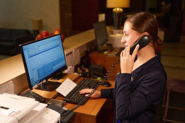 Side view photo of hotel administrator standing at reception counter while talking on phone and typing on computer clipart