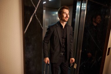 Elinde pasaport ve anahtar kartıyla asansörden inen gülümseyen adamın fotoğrafını çek.