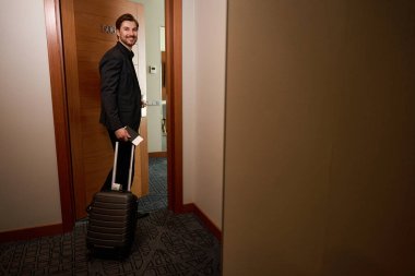 Takım elbiseli neşeli adam otel odasına elinde bavul ve pasaportla girer.