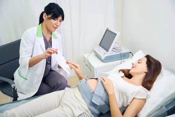 欧洲的成年女妇科医生在诊所向年轻的 面带微笑的白俄罗斯孕妇展示她腹部的心电图 怀孕检查的概念 — 图库照片