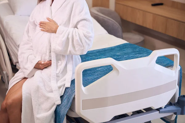 診療室の医療ソファーに腹を抱えているバスローブを着用している妊婦の一部 妊娠と出産の概念について — ストック写真