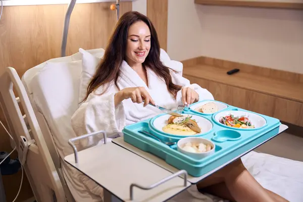 若い笑顔のヨーロッパの妊婦は 病院の部屋で医療ソファーで昼食をとっている間にナイフとフォークで肉を切るバスローブを身に着けています 妊娠と出産の概念について — ストック写真