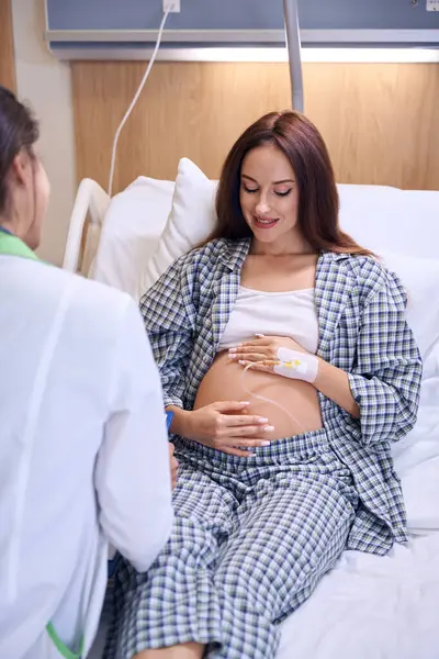 病院の医療ベッドで出産する前に 若い白血病の妊婦を調べる女性看護師の背中を閉じ込めました 妊娠と出産の概念について — ストック写真