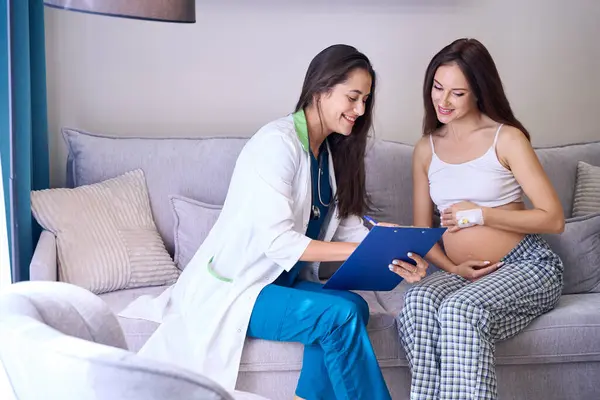 クリップボードを見ている若い白人妊婦は 診療所の医療室でソファーにヨーロッパの女性看護師を書いている 妊娠と出産の概念について — ストック写真