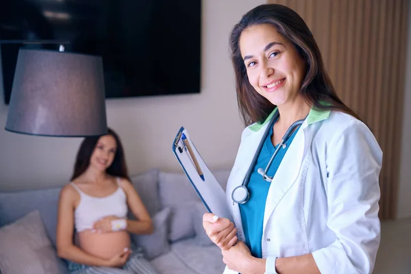 若い笑顔のヨーロッパの女性看護師の前景に焦点を当て クリップボードは診療所の医療室のソファーのぼやけた妊婦の背景を背景にカメラを見ています 妊娠と出産について — ストック写真
