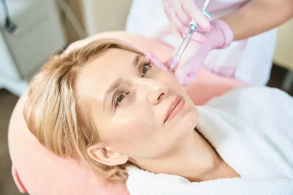 Partial female beautician doing face beauty injection with syringe of adult caucasian woman in blurred beauty salon. Face skin care, lifting and rejuvenation