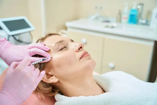 Cropped female beautician doing face beauty injection with syringe of adult caucasian woman with closed eyes in blurred beauty salon. Face skin care, lifting and rejuvenation