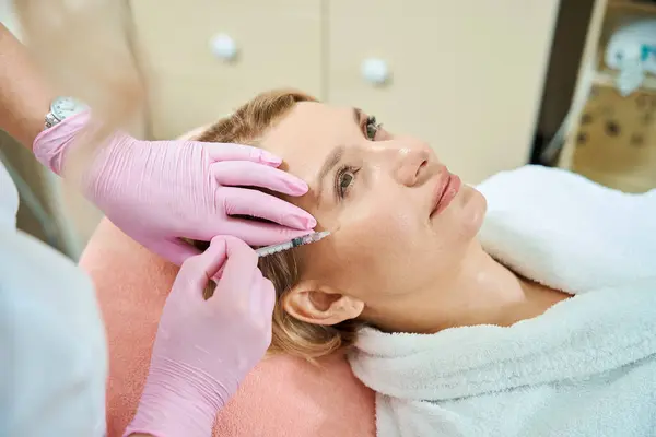 Cropped female beautician beautician doing face beauty injection with syringe of adult caucasian woman in blurred beauty salon. Face skin care, lifting and rejuvenation