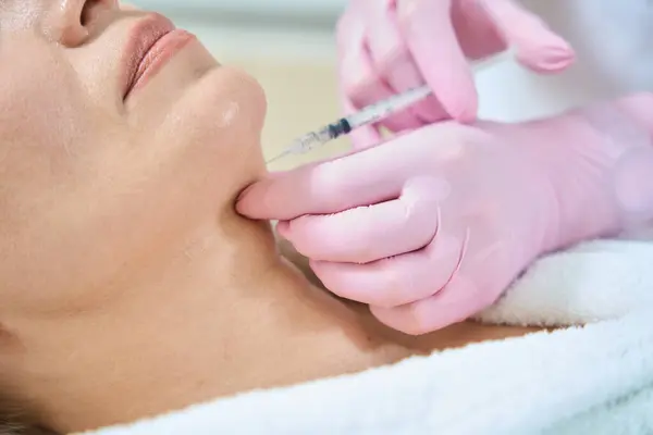 Cropped image and close up of female beautician doing face beauty injection with syringe of obscure face adult woman in blurred beauty salon. Face skin care, lifting and rejuvenation
