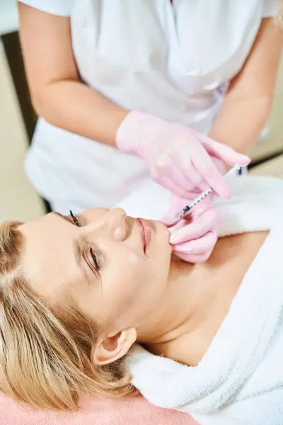 Partial female beautician doing face anti-aging injection with syringe of adult caucasian woman in blurred beauty salon. Face skin care, lifting and rejuvenation
