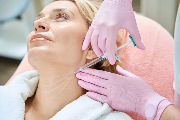 Cropped female beautician doing neck beauty injection with syringe of adult caucasian woman in blurred beauty salon. Body skin care, lifting and rejuvenation