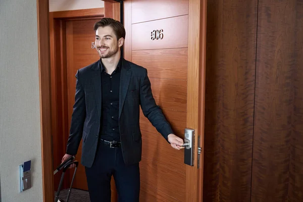 Young satisfied man in suit opens a door and goes into hotel room while holding trolley bag in hand