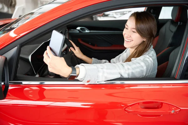 Cheerful woman sitting in car with mobile phone making selfie after buying new automobile in dealership