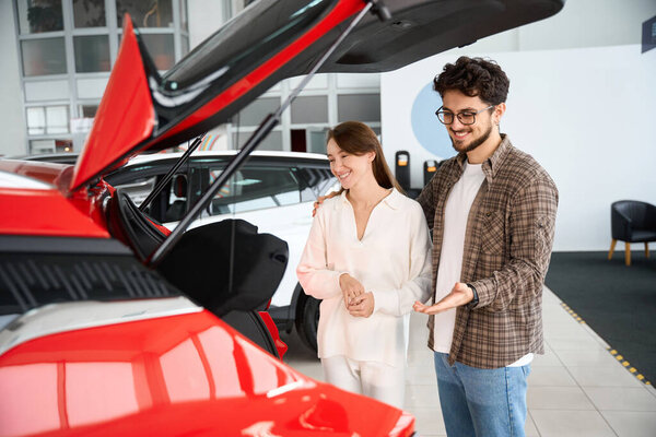 Привлекательная веселая женщина и мужчина стоят рядом с современным красным автомобилем и покупают новый автомобиль в автосалоне