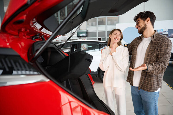 Счастливый радостный мужчина и женщина стоят у открытого багажника своего нового автомобиля в салоне и радуются покупке