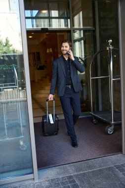 Takım elbiseli neşeli adam elinde bavulla otelden çıkarken telefonla konuşuyor.