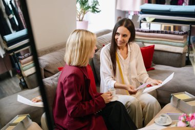 Tasarım stüdyosunda iki kadın skeçlere bakıyor, kadınlar rahat bir kanepede oturuyor.