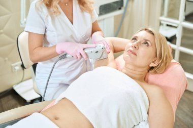 Kısmi kadın güzellik uzmanı, bulanık güzellik salonundaki yetişkin beyaz bir kadının lazer tüy temizleme makinesiyle lazer tüy temizleme yapıyor. Vücut bakımı kavramı