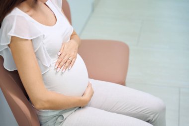 Klinikte konsültasyon beklerken sandalyeye oturup karnına dokunan hamile bir kadının kısmi görüntüsü. Hamilelik testi kavramı