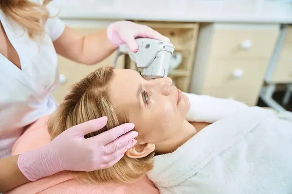 Partial female beautician doing laser depilation with laser hair removal machine of forehead of adult caucasian woman wearing bathrobe in blurred beauty salon. Concept of face skin care