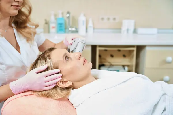Obscure face of female cosmetologist doing laser depilation with laser hair removal machine of face of adult caucasian woman in blurred beauty salon. Concept of face skin care