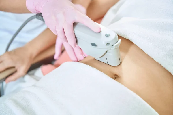 Cropped image of female beautician doing laser depilation with IPL photorejuvenation device of woman belly in blurred beauty salon. Concept of body skin care
