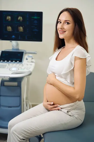 Piękna Uśmiechnięta Biała Kobieta Ciąży Patrząca Kamerę Siedzącą Medycznej Kanapie — Zdjęcie stockowe