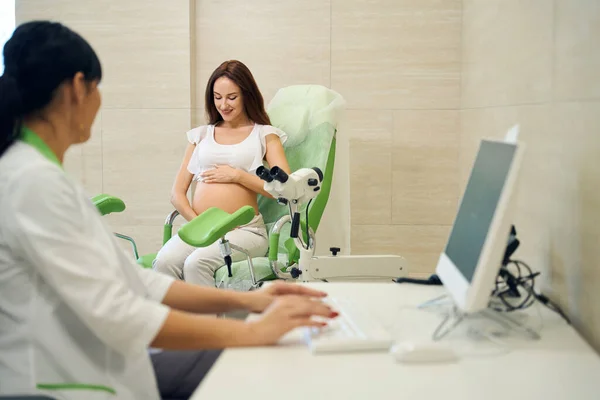 女妇科医生在电脑上打字 在诊所检查时看着年轻的 面带微笑的高加索孕妇看着她的腹部 怀孕检查的概念 — 图库照片