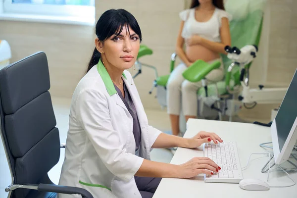 欧洲成年女妇科医生一边在电脑上打字 一边看着相机 并在诊所的医疗沙发上检查模糊的孕妇 怀孕检查的概念 — 图库照片