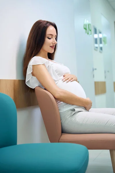 若い白人妊婦が椅子に座り 診療所のホールで診察を待っている間に腹を見つめていた 妊娠検査の概念 — ストック写真