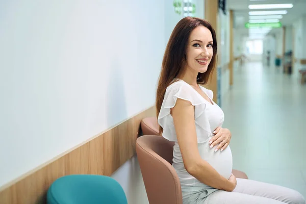 若い笑顔のヨーロッパの妊婦が椅子に座り 診療所のホールで相談を待っている間にカメラを見ている 妊娠検査の概念 — ストック写真