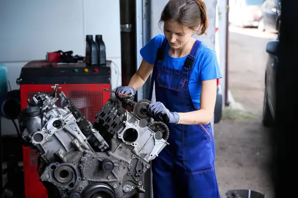 Woman is repairing a large auto part in an auto repair shop, she is wearing a blue uniform